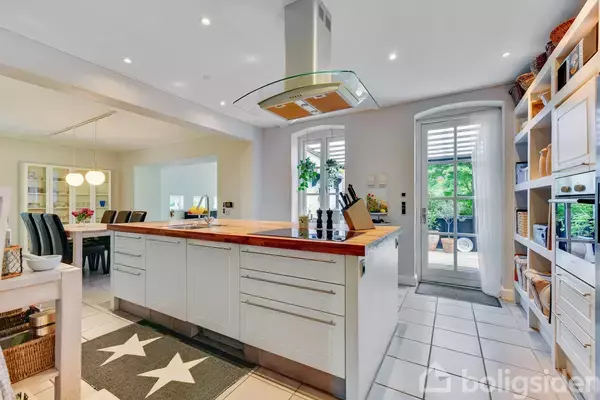 Et moderne køkken med en central ø i træ, overdækket af en emhætte. Omkring er der hvide skabe, hylder med dekorationer, og en døråbning til en terrasse med grøn udsigt.