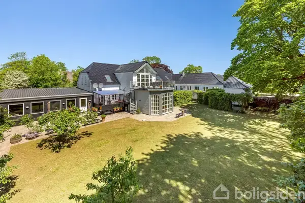 Et stort, hvidt hus med flere sektioner og en stor have omgivet af grønne træer under en klar blå himmel.