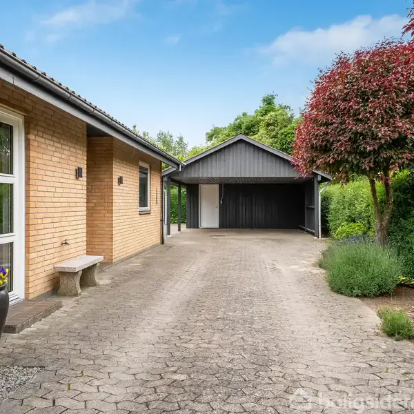 Huset har gule mursten og står ved siden af en flisebelagt indkørsel, der fører til en garage. Omkring er der frodig beplantning og en rødbladet busk ved siden af plænen.