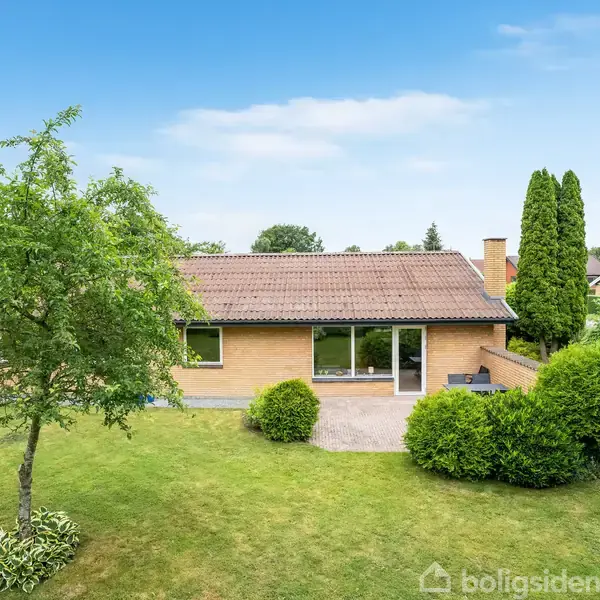 En gul murstensbungalow med brun tagsten ligger stille i en velplejet have med grønne buske og et træ. Huset har en stor glasdør mod terrassen under en blå himmel.