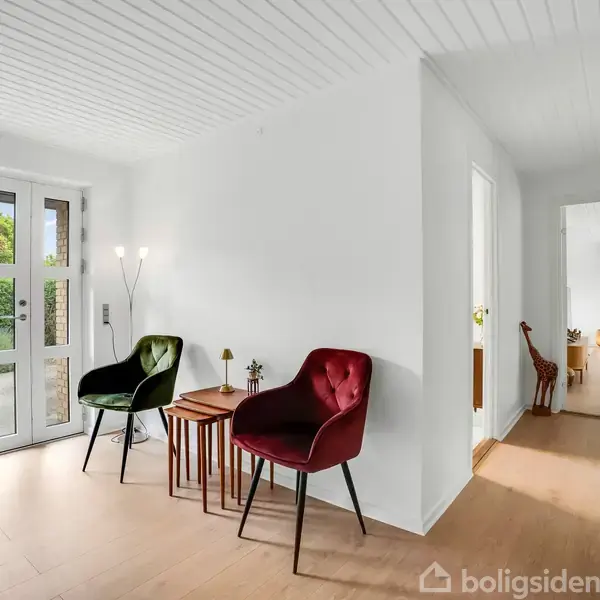 To stole, en grøn og en rød, står ved et lille bord i en indgang med hvide vægge. En dør og et vindue lader lys ind. En gang leder videre.