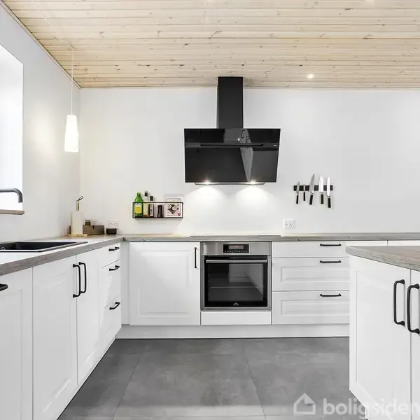 Hvidt køkken med sorte greb på skabe. Integreret ovn under sort emhætte. Lys strømmer ind gennem vinduet til venstre. Lys træloft og gråt gulv. To lys hænger ved siden af vasken.