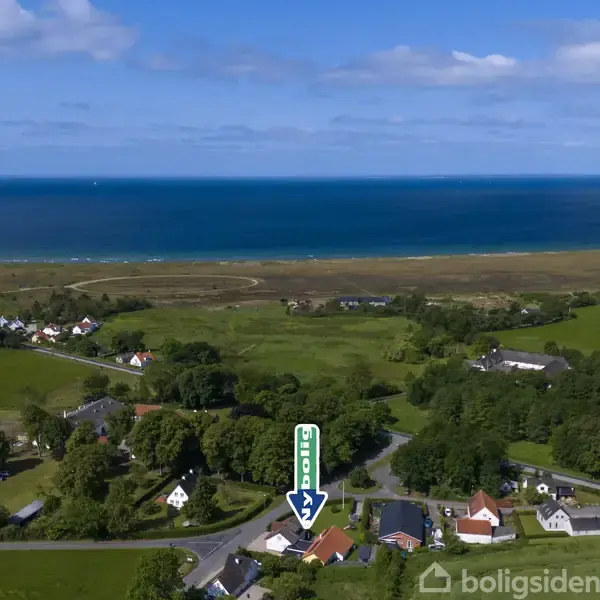En pil peger på et rødt hus, omgivet af grønne marker og flere huse. I baggrunden ses kystlinje og hav. Pilens tekst: "Bolig."