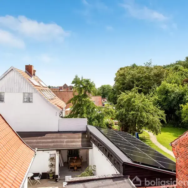 Røde tegltagshuse omgiver en gårdsplads med et lille siddeområde. Et sort tag på et skur har solpaneler. I baggrunden ses en frodig have med træer under en klar blå himmel.