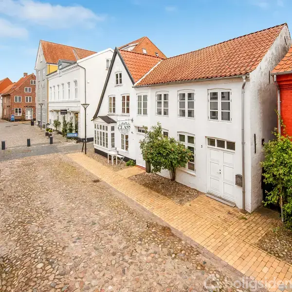 Gader med brosten og række af charmerende huse; hvide facader og et enkelt rødt hus med teglstenstage. Himlen er blå, og der er ingen synlig tekst.