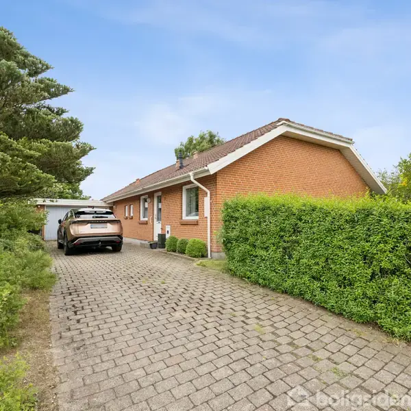 Et rødt murstenshus med et skråt tag ligger ved siden af en brolagt indkørsel. En bil holder parkeret i carporten, omgivet af frodig grøn beplantning.