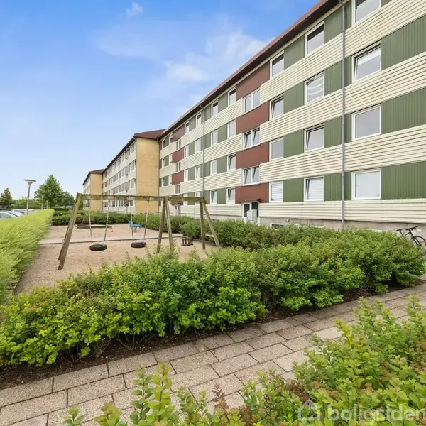 En stor bygning med farverige paneler står ved siden af en legeplads med gynger. Omgivet af grønne buske og en flisebelagt sti i et boligområde.