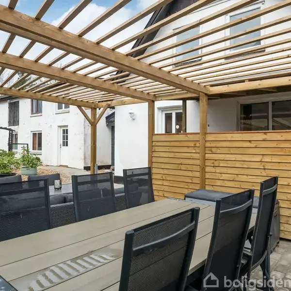 En lang udendørs træborde står under en pergola med stole omkring. I baggrunden ses et hvidt hus med brune vindusrammer og en gårdhave med planter.