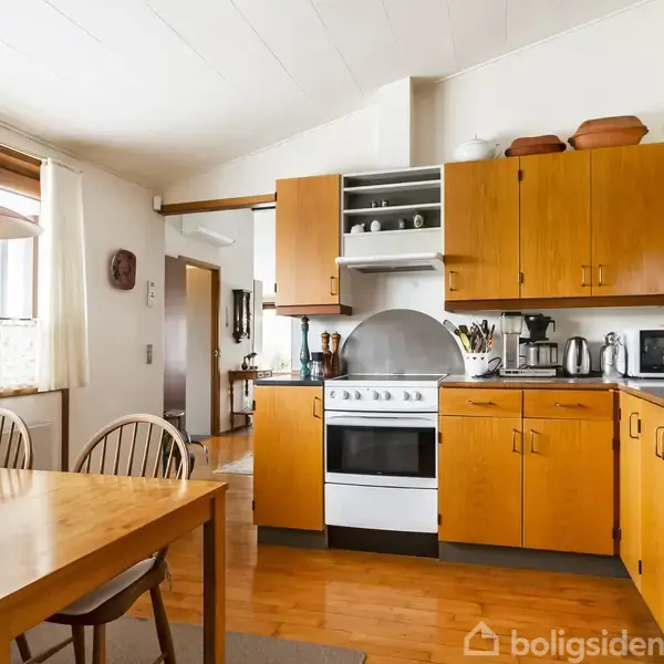 Et køkken med orange træskabe og hvidt komfur i midten. På bordet står forskellige køkkenredskaber, og en rød lampe hænger over et spisebord ved vinduet.