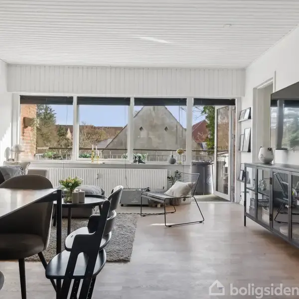 Stue med spisebord og stole til venstre, sofa og lænestole ved vindue, stort tv på højre væg. Lyse vægge og trægulv. Store vinduer med udsigt til have.