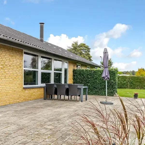 Gul murstenshus med terrasse, hvor fire sorte stole og et bord står ved siden af en sammenklappet parasol. Græsplæne og hæk omringer terrassen, og himlen er blå med skyer.