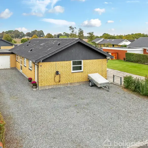 Et gult murstenshus med sort tag står i en forstad med grønne haver. En trailer er parkeret på en grusbelagt indkørsel foran huset.