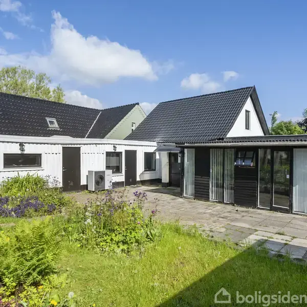 Et hvidt hus med sort tag står omgivet af en terrasse og have. Huset har store vinduer og en græsplæne med blomster og buske under en klar blå himmel.