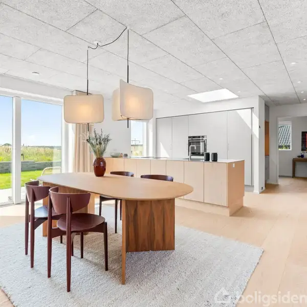 Et moderne køkken-alrum med et ovalt træbord og seks stole på et lyst tæppe. Store vinduer giver udsigt til en have. Lyse skabsmoduler og en ø oplyser rummet.