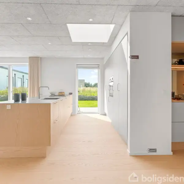 Et moderne køkken med lyst trægulv indeholder en køkkenø og skab. I baggrunden ses en spiseplads med stole og et stort vindue med udsigt til en grøn have.