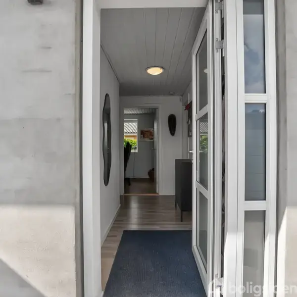 Dør står åben til en lys gang med lyst trægulv, en blåligt tæppe og vægpynt. En lampe lyser loftet, og en dør åbner til et rum for enden.