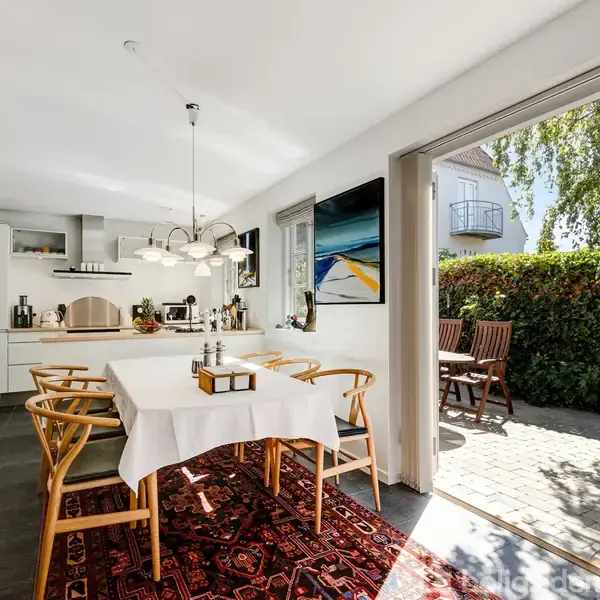 Et moderne, lyst køkken med et spisebord dækket med en hvid dug. Stole står omkring bordet, og en åben dør fører til en flisebelagt gårdhave omkranset af grønne hække.