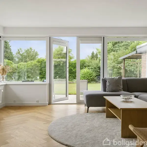 Grå sofa med puder står ved siden af et træbord i en lys stue. Døre åbner mod en grøn have. Store vinduer lader lys ind, med udsigt til træer.