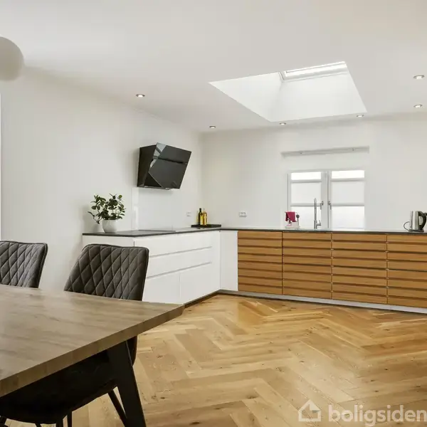 Et moderne køkken-alrum med en stor spisebord i forgrunden, flotte trægulve, hvide skabselementer og en skrå loftshal med ovenlysvindue. Vinduet slipper naturligt lys ind i rummet.