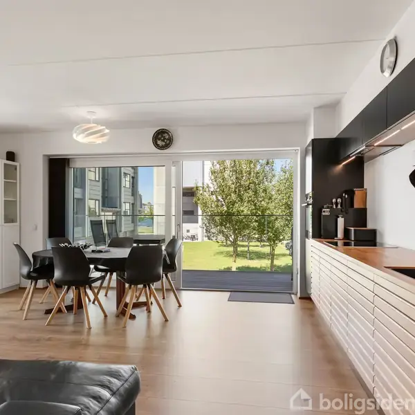 Moderne køkken-alrum med trægulve og mørke møbler. Et spisebord med stole står ved en stor glasdør, der åbner mod en have. Det indbyggede køkken er stilfuldt med træpaneler.