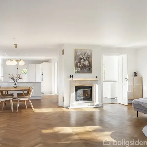 En lys stue med parketgulv har en pejs i midten, et spisebord til venstre og en sofa til højre. Køkken ses i baggrunden. Ingen tekst i billedet.