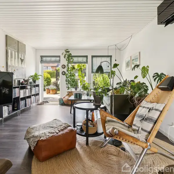 Stue med lænestol og tv, omgivet af grønne planter og bøger. Store vinduer tillader naturligt lys at strømme ind. Møbler og interiør skaber en hyggelig atmosfære.
