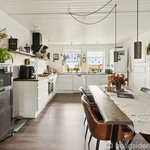Køkken med lang spisebord, omgivet af brune stole. Bordet er dekoreret med en lys løber og en blomsteropsats. Køkkenet har hvide skabe, hængende lamper og vinduer med udsigt til planter udenfor.