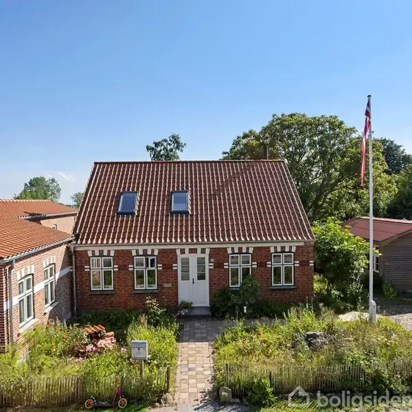 Et rødt murstenshus med rødt tegltag står i en have med blomsterbede og en flagstang. På venstre side ses en tilstødende bygning, og i baggrunden er der træer.