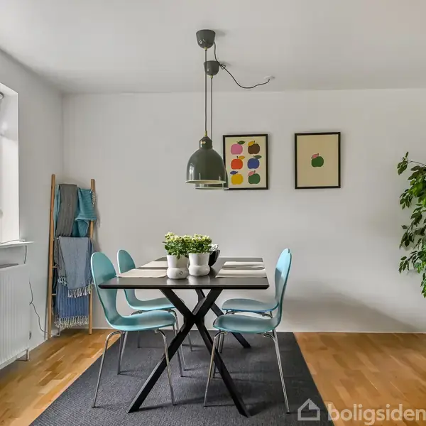 Spisebord med fire blå stole står på et tæppe i et lyst rum. Over bordet hænger en lampe, og ved siden af er en stor plante. Væggen har to billeder.