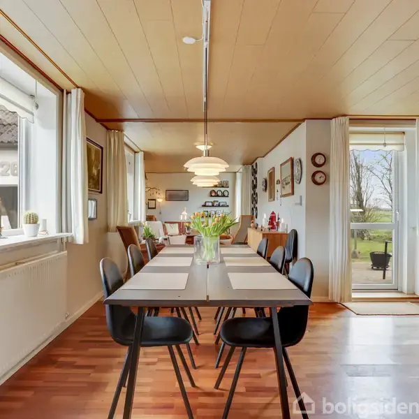 Langt spisebord med sorte stole står dækket i et lyst køkken-alrum. Rummet har store vinduer med udsyn til en grøn have. Der hænger moderne lamper fra loftet.
