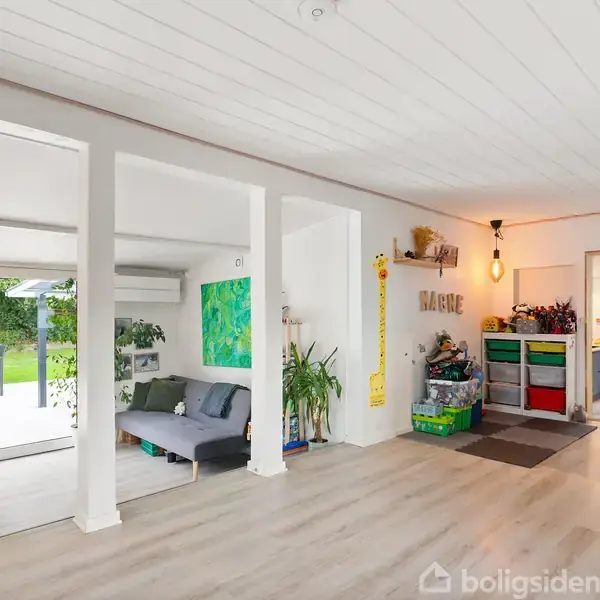 Stue med trægulv og lys indretning, sofa ved glasdøre ud til have. Maleri og planter skaber hygge. Åben overgang til køkken med moderne møblering. Børnereoler og lys dekoration.