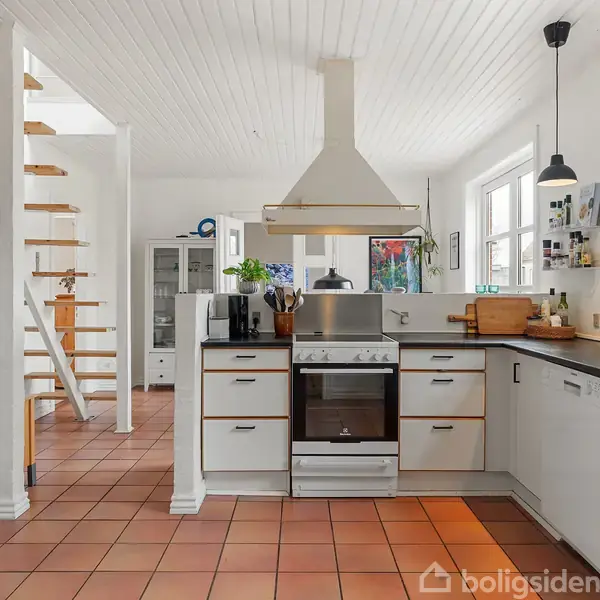Spisebord med fire sorte stole i moderne køkken-alrum. Bordet er dækket med dækkeservietter, blomsterbuketter, og lysestager. Rummet er lyst med store vinduer og åben trappe i baggrunden.