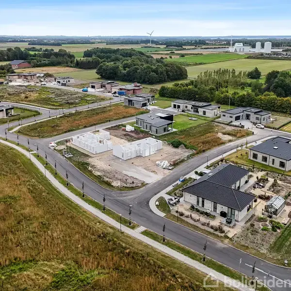 Nybyggeri i et boligområde med flere moderne huse under konstruktion. Omkringliggende natur med marker og spredte træer i baggrunden. Asfalterede veje forbinder ejendommene i bebyggelsen.