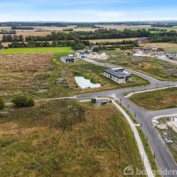 En mark ligger i et åbent område med få moderne huse placeret i byggegrunde. En smal vej og en lille dam vises blandt de grønne områder, marker og træer.