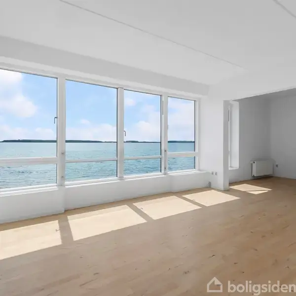 Et lyst og tomt værelse med trægulv har store vinduer, der giver udsigt over havet. Rummet er minimalistisk indrettet med en radiator langs væggen.