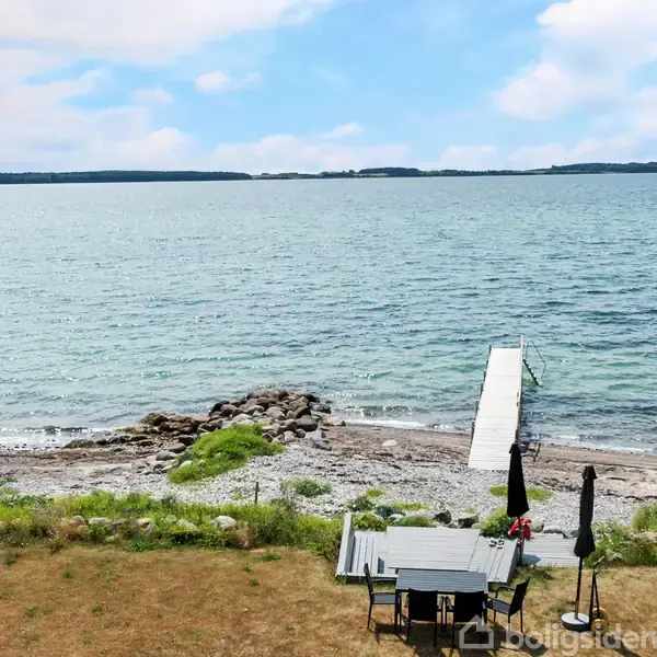 Et havesæt står stille på en græsplæne tæt ved en stenstrand med udsigt over et bredt, roligt vand og en lang, smal bådebro, omgivet af lette skyer og horisont.
