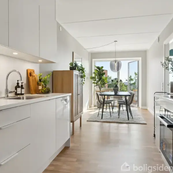 Moderne køkken med hvide skabe, stålvask, og træbordplade; spisebord med stole står ved vinduerne. Flere grønne planter skaber en frisk atmosfære i det lyse rum.