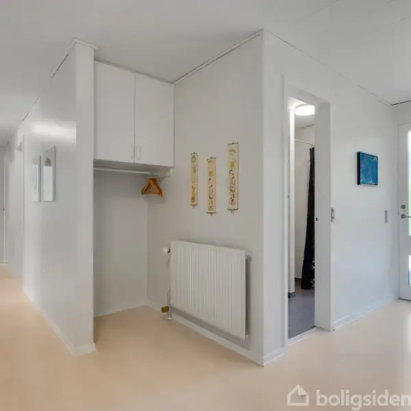 Indgangsparti med bøjleholder og radiator, lyst interiør, døre til forskellige rum. Dæmpet hvid belysning og indrammede billeder på væggene. Døren fører til en udendørs scene.
