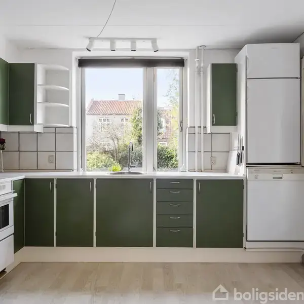 Grønt køkken med hvide detaljer, uden aktivitet. Rummet har indbygget skab, komfur, og et køleskab. Store vinduer med udsigt til en have. Lyst trægulv og flisebelægning på væggene.