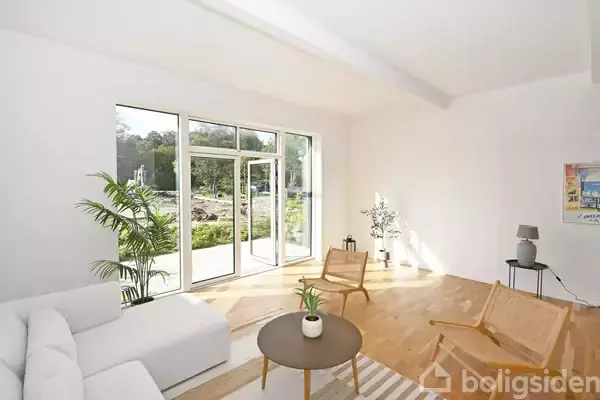 En lys stue med en hvid sofa og to træstole omkring et rundt sofabord. Store vinduer åbner ud til en grøn have. En moderne og minimalistisk indretning.