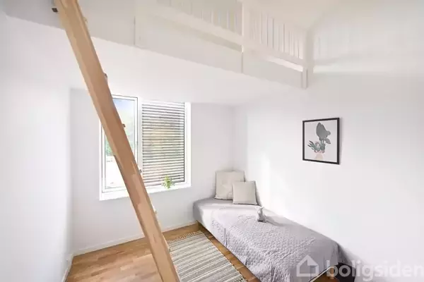 En stige leder op til en hems i et lyst rum med en seng, tæppe og vægkunst. Et vindue slipper naturligt lys ind, og gulvet er af træ.