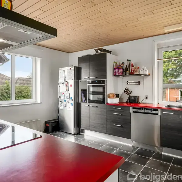 Et moderne køkken med sorte skabe og røde bordplader. Køleskabet har noter og magneter. Store vinduer giver udsigt til et grønt område. Flisegulv og træloft.