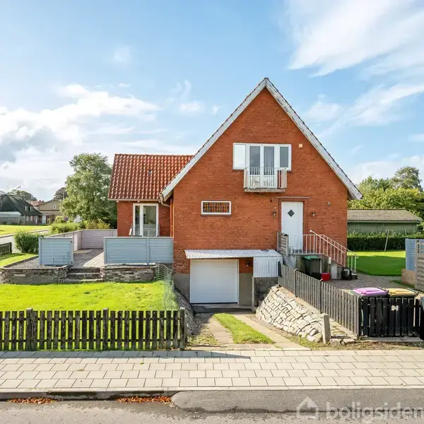 Rødt murstenshus står stille omgivet af en grøn græsplæne og hvid stakit. Taget er rødt, og en trappe fører til indgangen. En garage ligger nedenunder.