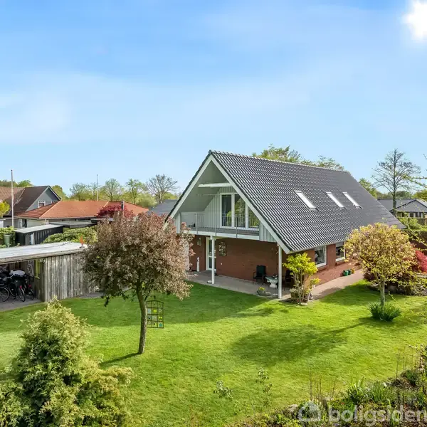 Et stort hus med skråtag står i en velplejet have. En carport er til venstre, og flere træer omgiver ejendommen i et beboelseskvarter.