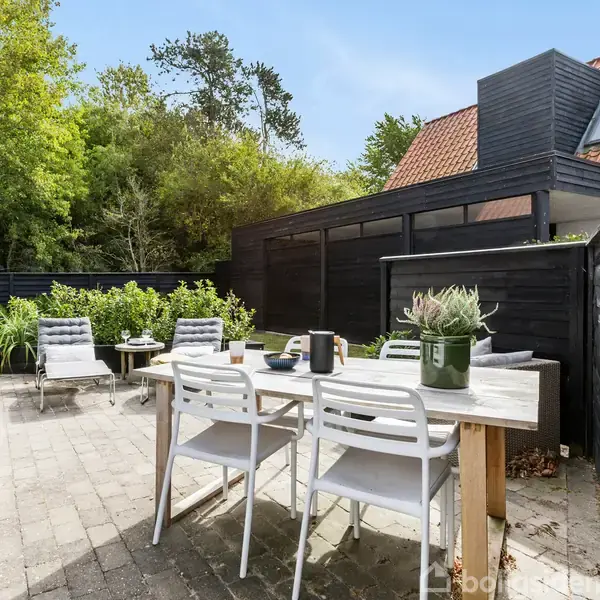 En terrasse med et hvidt bord og fire stole, hvorpå der står en plante og diverse genstande. Omgivet af et sort plankeværk, med træer i baggrunden under en blå himmel.
