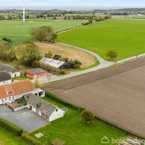 Et hvidt hus med rødt tag står stille omgivet af en stor gårdsplads og grønne marker. Baggrunden viser åbne marker, en grusvej, og en vindmølle.