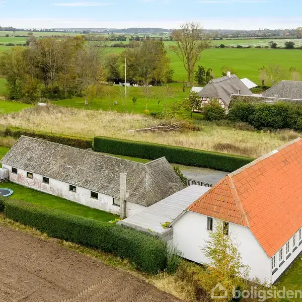 Hvidt bondehus med rødt tag ligger blandt marker og grønne områder. Ved siden af står et længere stråtagbygning. Baggrunden domineres af åbne marker og spredte træer under en blå himmel.
