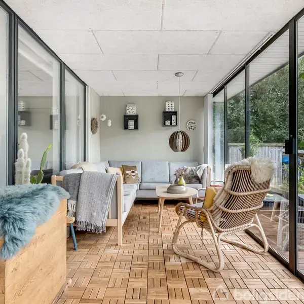 Stue med glasvægge, hvor en lys sofa står mod bagvæggen. En flettet stol er placeret foran og et grønt træ ses udenfor. Trægulve og tæpper tilføjer varme.