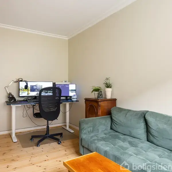 En kontorstol står foran et skrivebord med to skærme i en stue med trægulv. Til højre er en grøn sofa ved siden af en lille trækommode med planter.