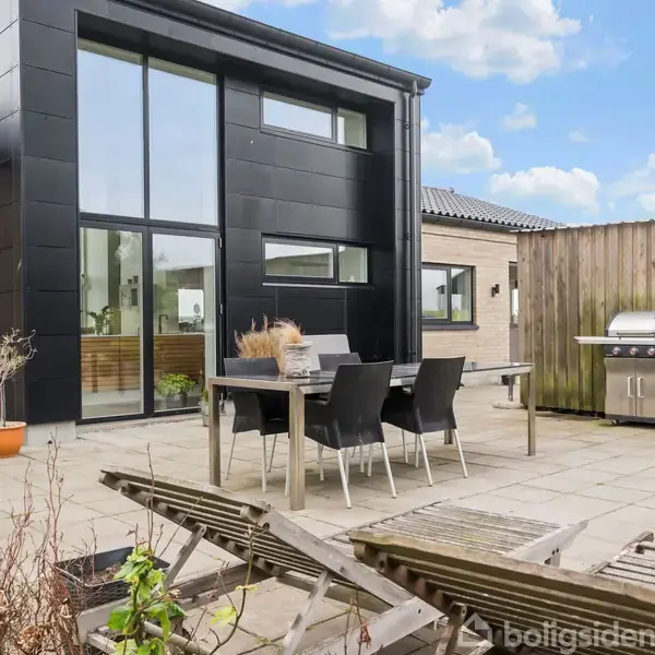 Et moderne hus med store vinduer har møbleret terrasse. På terrassen står et spisebord med stole og en grill ved træhegn. Haven har potteplanter og liggestole.